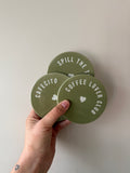 Drie ronde olijfgroene onderzetters met witte teksten Coffee Lover Club, Cafecito en Spill the Tea