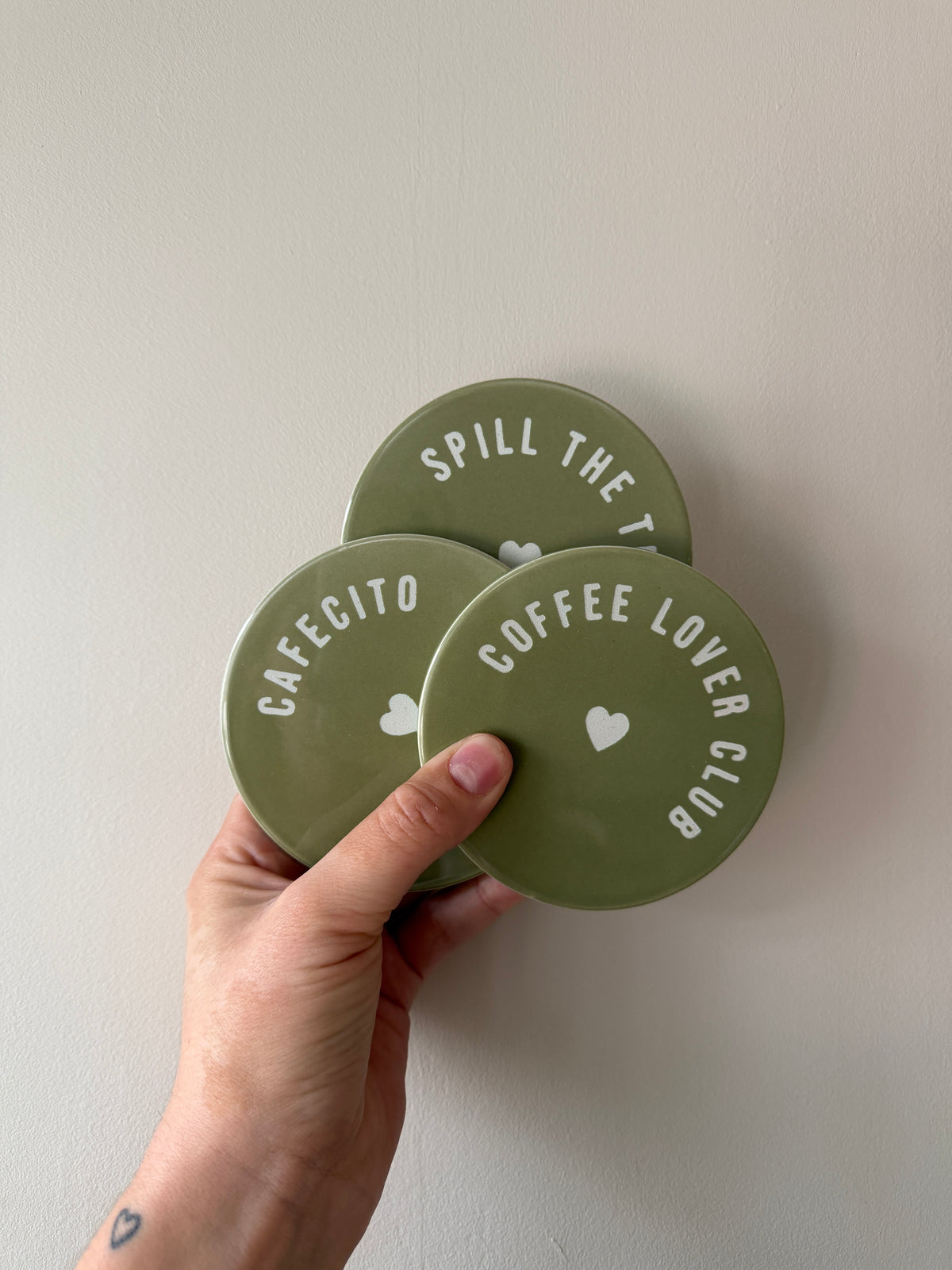 Drie ronde olijfgroene onderzetters met witte teksten Coffee Lover Club, Cafecito en Spill the Tea